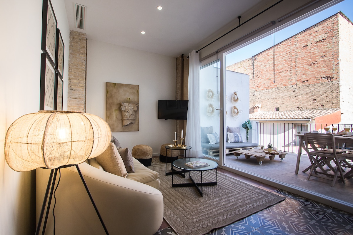 A light-filled living area showcases a comfortable beige sofa and a woven area rug. A modern lamp stands beside the couch, while a TV is mounted on the wall. Large sliding glass doors open to a balcony with outdoor seating, enhancing the spacious ambiance.
