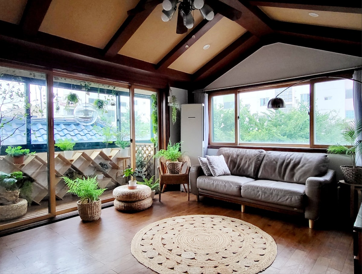 A bright living area features large windows adorned with greenery, allowing natural light to fill the space. A comfortable gray sofa is positioned beside a round, woven rug. Decorative plants and a small table enhance the cozy atmosphere, creating a welcoming environment for relaxation.
