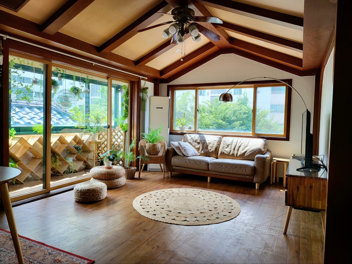 A well-lit living area features a comfortable sofa arrangement beside large windows, showcasing views of greenery outside. A round rug lies on the wooden floor, complemented by a floor lamp and decorative poufs. Natural light streams in, highlighting the wooden beams of the ceiling.