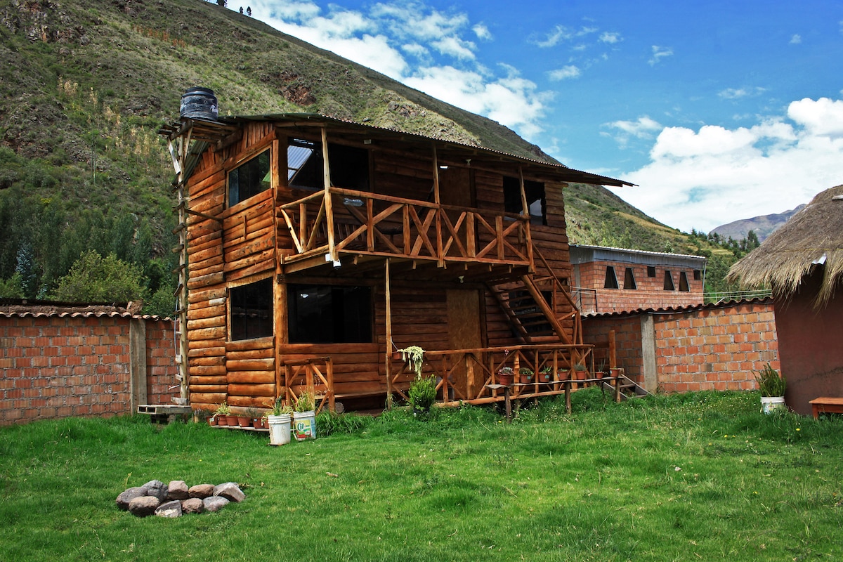 A wooden cabin structure is set amidst lush green grass, flanked by a mountain backdrop. A staircase leads to a second floor balcony, which is adorned with wooden railings. Surrounding vegetation and a stone fire pit enhance the natural setting.