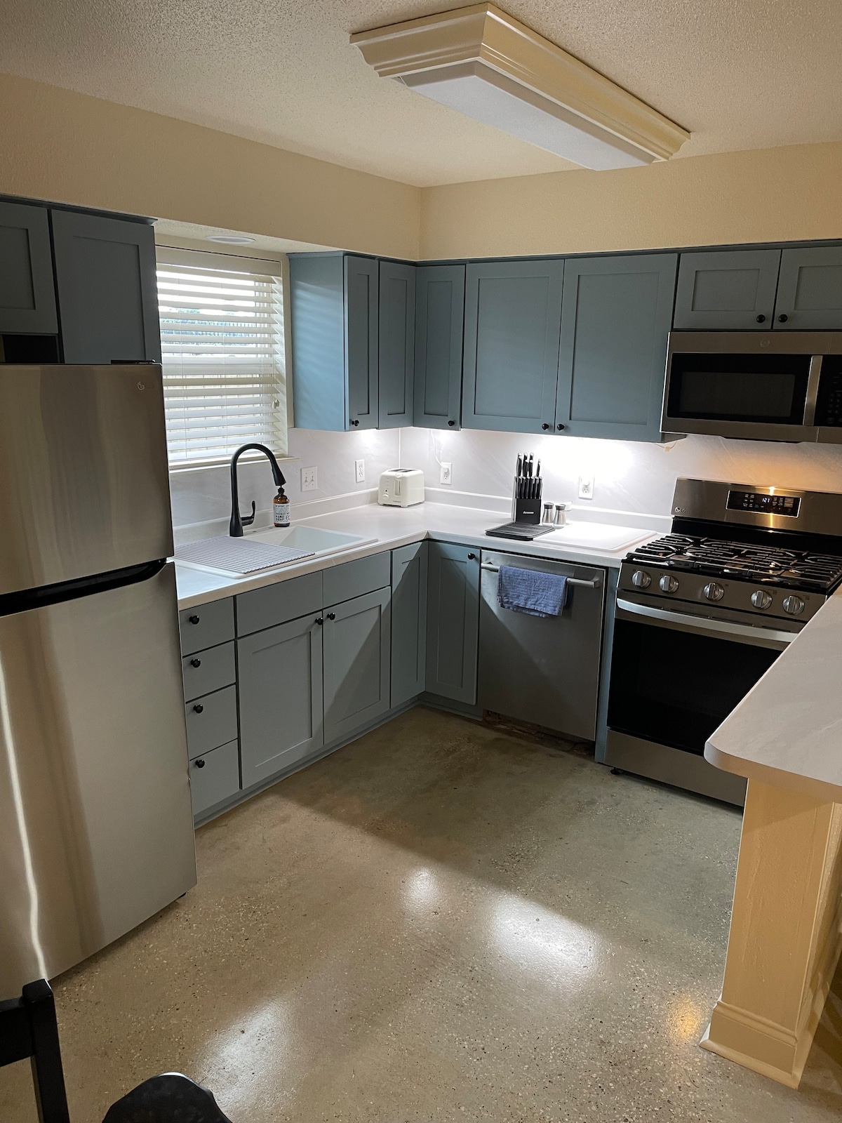 The kitchen features light blue cabinetry, stainless steel appliances, and a neutral countertop. A window provides natural light, and modern fixtures, including a sleek faucet and a gas stove, enhance the functionality. A small area for essential kitchen tools is also visible.