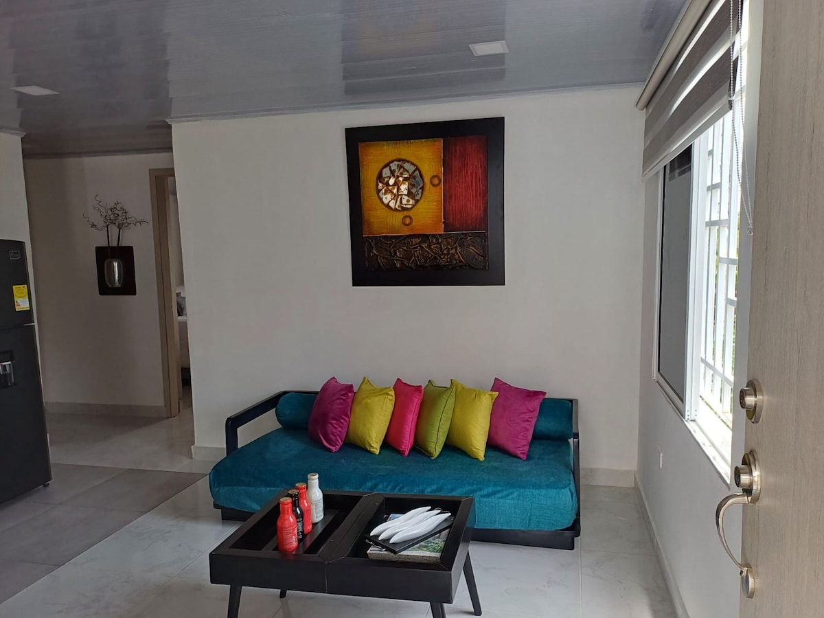 A comfortable seating area features a sleek blue couch adorned with colorful pillows in vibrant shades of pink and green. A low coffee table sits in front, displaying various items. Bright natural light streams in through a large window, complemented by elegant wall art.