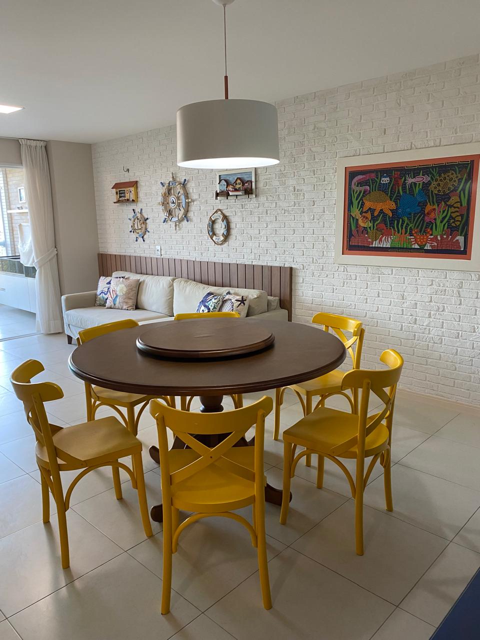 A dining area is highlighted by a round wooden table surrounded by six yellow chairs. A light fixture hangs above the table, while a soft sofa with decorative pillows is positioned against a textured wall. Artwork featuring vibrant colors is displayed nearby.
