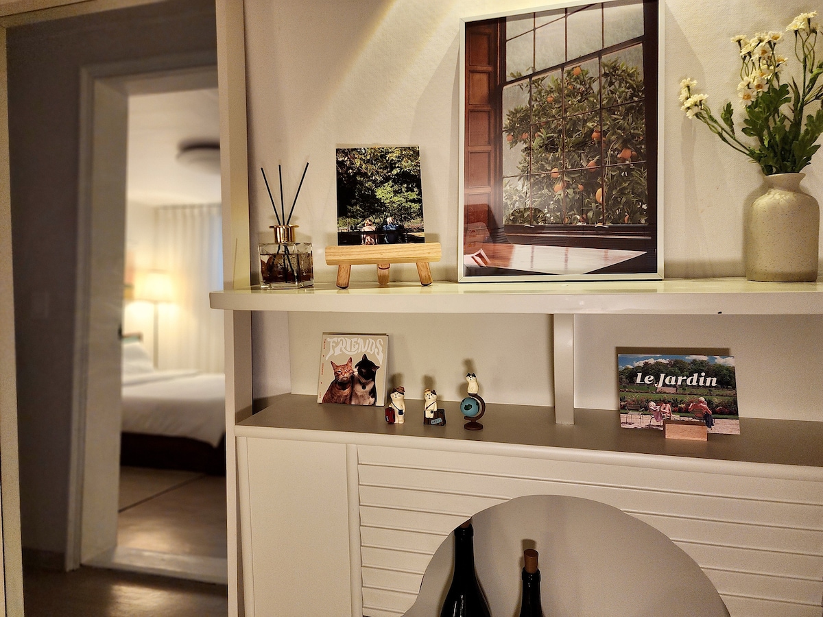 A neatly arranged shelf displays framed photos and small decorative items, including a vase with fresh flowers. Dim lighting creates a cozy atmosphere while soft textures and colors invite tranquility. A glimpse of the bedroom can be seen in the background.