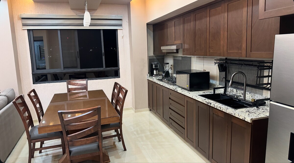 A modern kitchen and dining area are shown, featuring dark wood cabinetry and stainless steel appliances. A dining table with six wooden chairs occupies the space, while a black sink sits under a window. A light fixture hangs above, contributing to the overall ambiance.