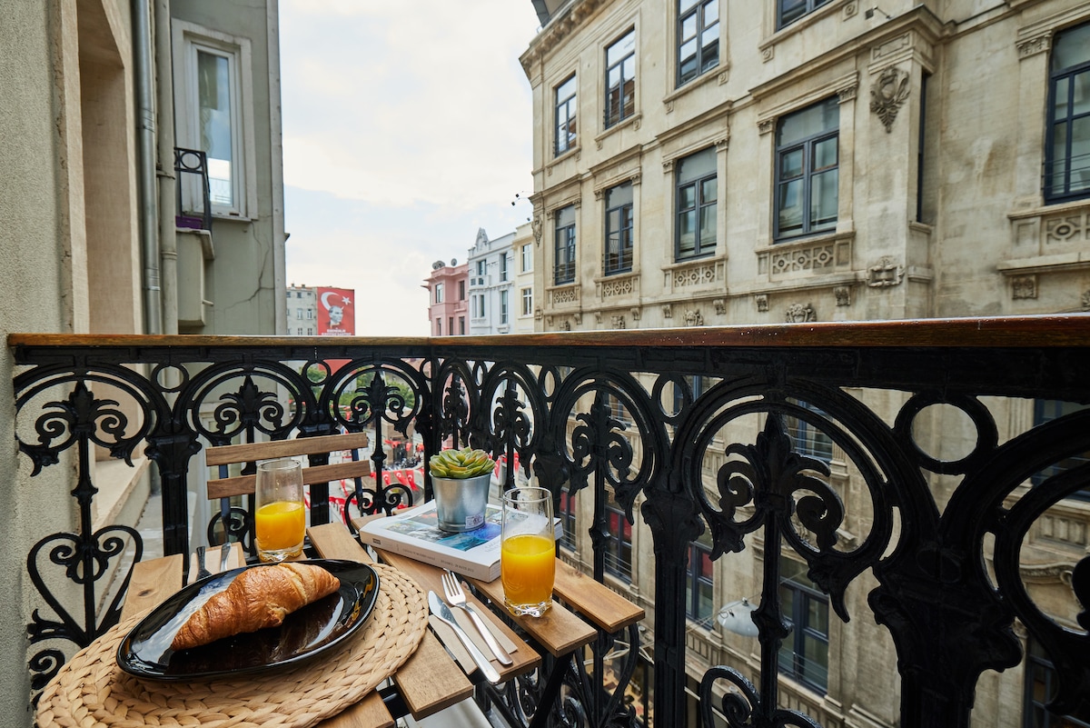 A balcony with a decorative wrought iron railing displays a small wooden table set for breakfast. A croissant, two glasses of orange juice, and a succulent plant are arranged on the table. The backdrop features historic buildings lining the bustling street.