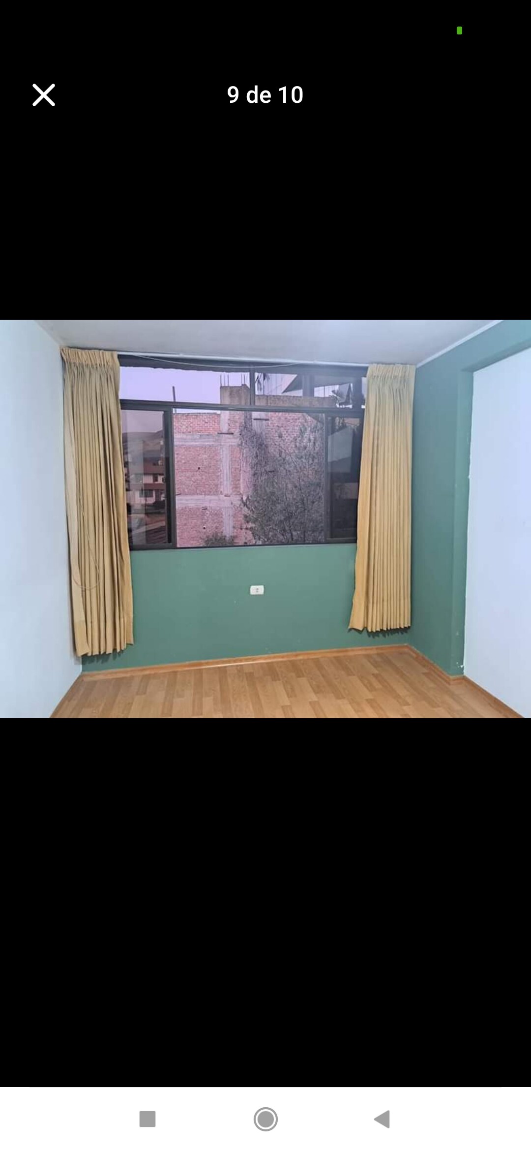 A corner room is presented with a large window framed by golden curtains, allowing natural light to enter. The walls are painted in a calming green hue, complemented by a polished wooden floor, providing a simple and inviting space.