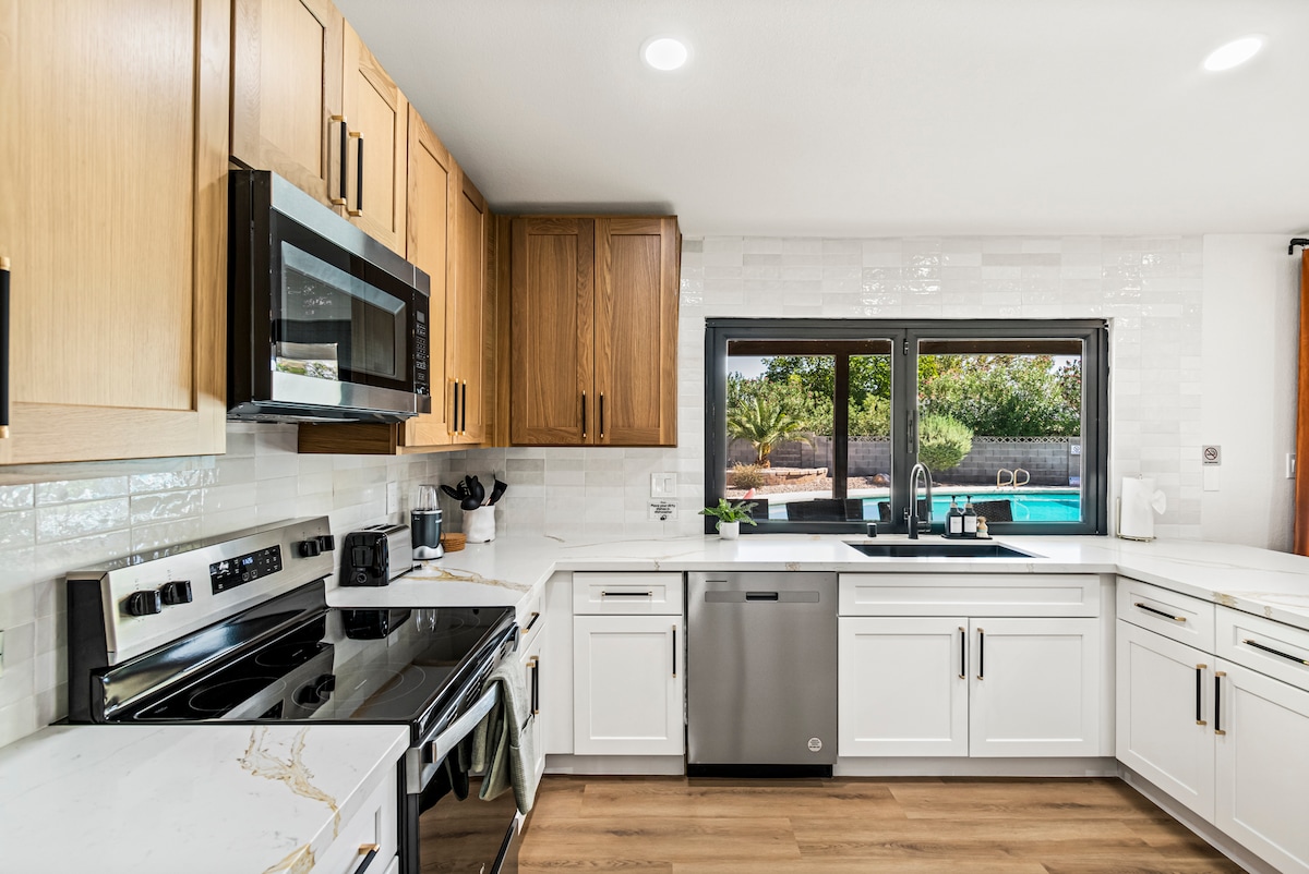 The kitchen showcases modern cabinetry in contrasting light and dark wood tones. Stainless steel appliances, including a microwave and oven, are visible. A large window offers a view of the outdoor space, allowing natural light to illuminate the counter surfaces.