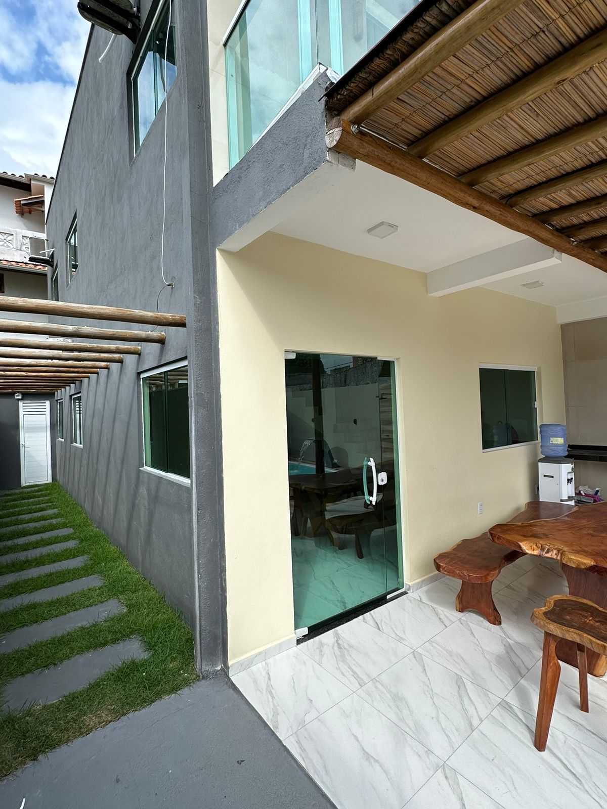 An entrance featuring large glass doors is shown, providing a view into the interior. The side of the building is complemented by a well-maintained garden path of stone tiles and grass, leading towards an outdoor dining area, visualized under a shaded roof.