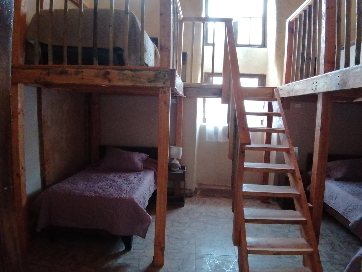 A shared room featuring wooden bunk beds and single beds. Natural light enters through large windows, illuminating the space. A staircase leads to the upper bunk, and bedside tables with lamps are positioned near the lower beds, creating a functional layout.