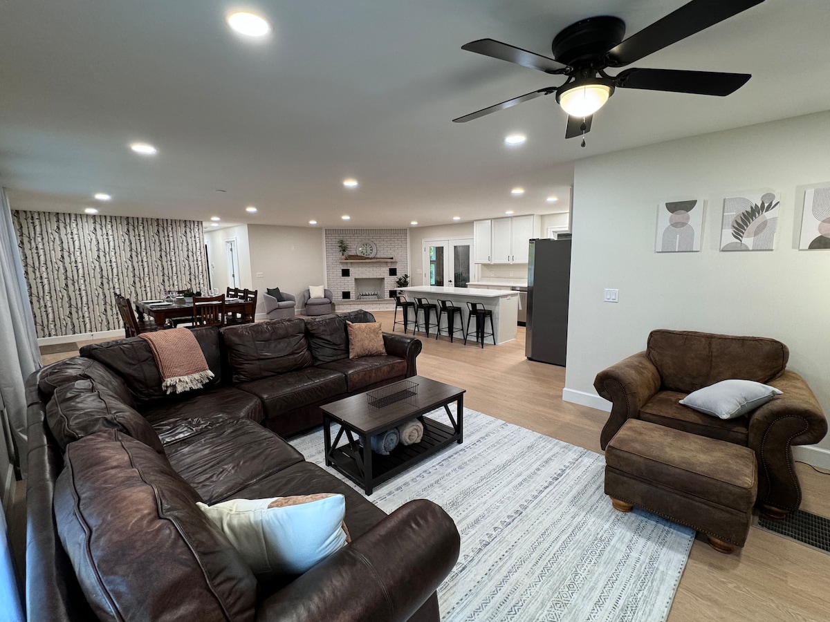 A spacious living room features a large sectional sofa arranged around a coffee table, with additional seating provided by a plush armchair. Modern light fixtures illuminate the room. The decor includes light-colored walls, a patterned area rug, and soft window treatments that enhance the comfort of the space.