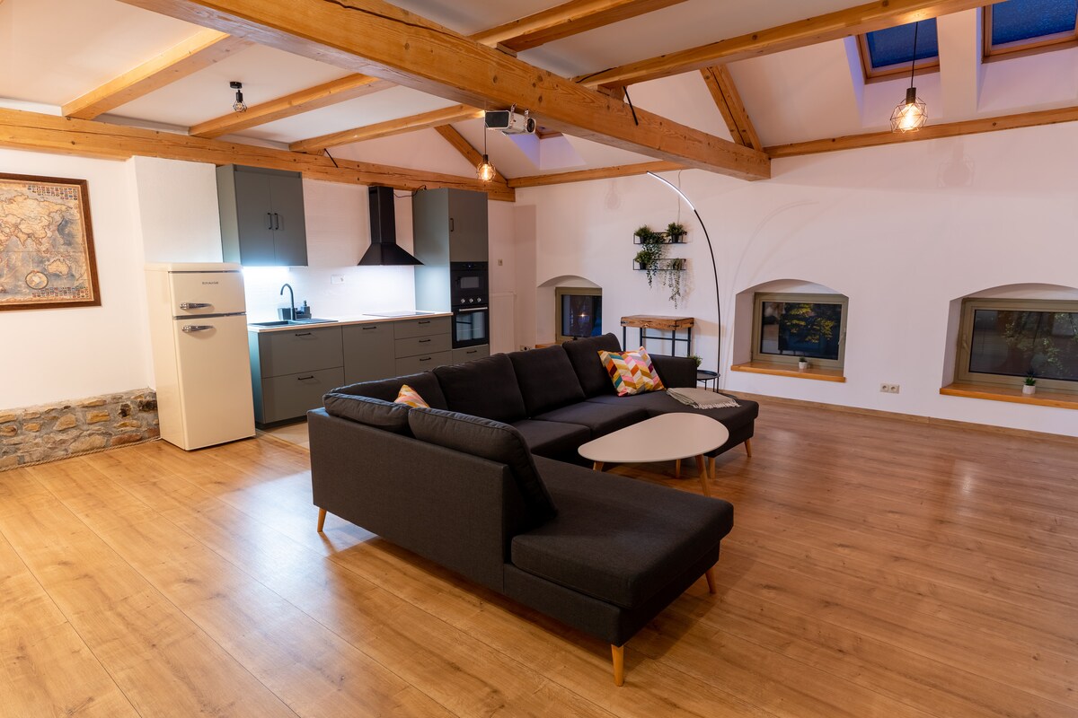A spacious living area is highlighted by wooden beams and warm lighting. A large sectional sofa is positioned next to a modern coffee table. The kitchen area is visible, featuring sleek cabinetry and appliances. Natural light enters through several window niches, enhancing the space.