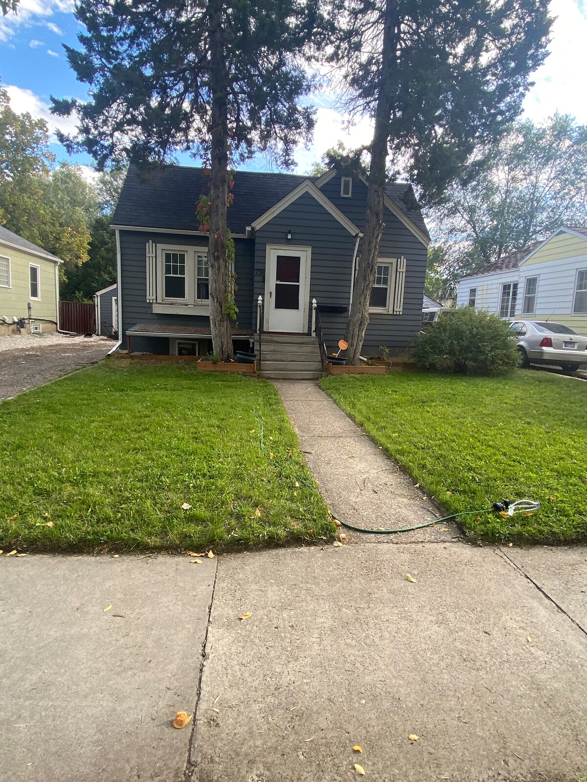 The exterior of the retreat is shown, featuring a blue-gray house with a peaked roof surrounded by well-maintained grass. A concrete pathway leads to the front door, flanked by trees. Parking spaces are visible nearby, indicating accessibility.