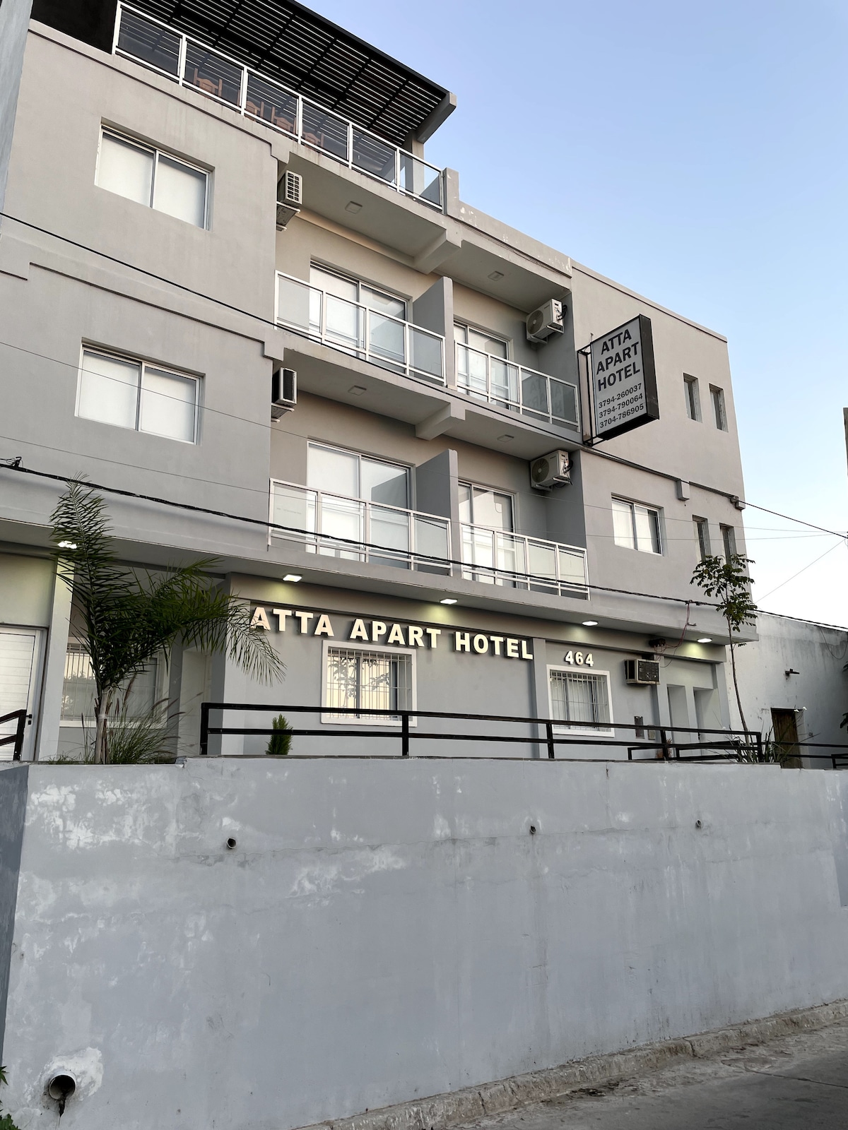 The exterior of Atta Apart Hotel features a modern design with multiple balconies on the upper floors. The building is painted in light gray, with the hotel name prominently displayed. A small garden area is located at the front, enhancing the inviting appearance.