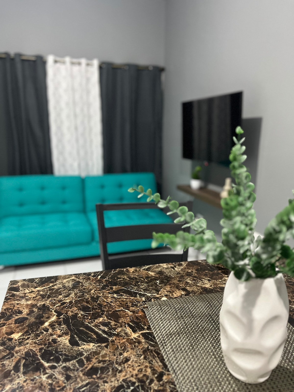 A dining area is highlighted by a dark marble table accented with a vase containing greenery. A turquoise sofa is visible in the background, accompanied by dark curtains and a wall-mounted television, creating a cohesive and stylish living space.