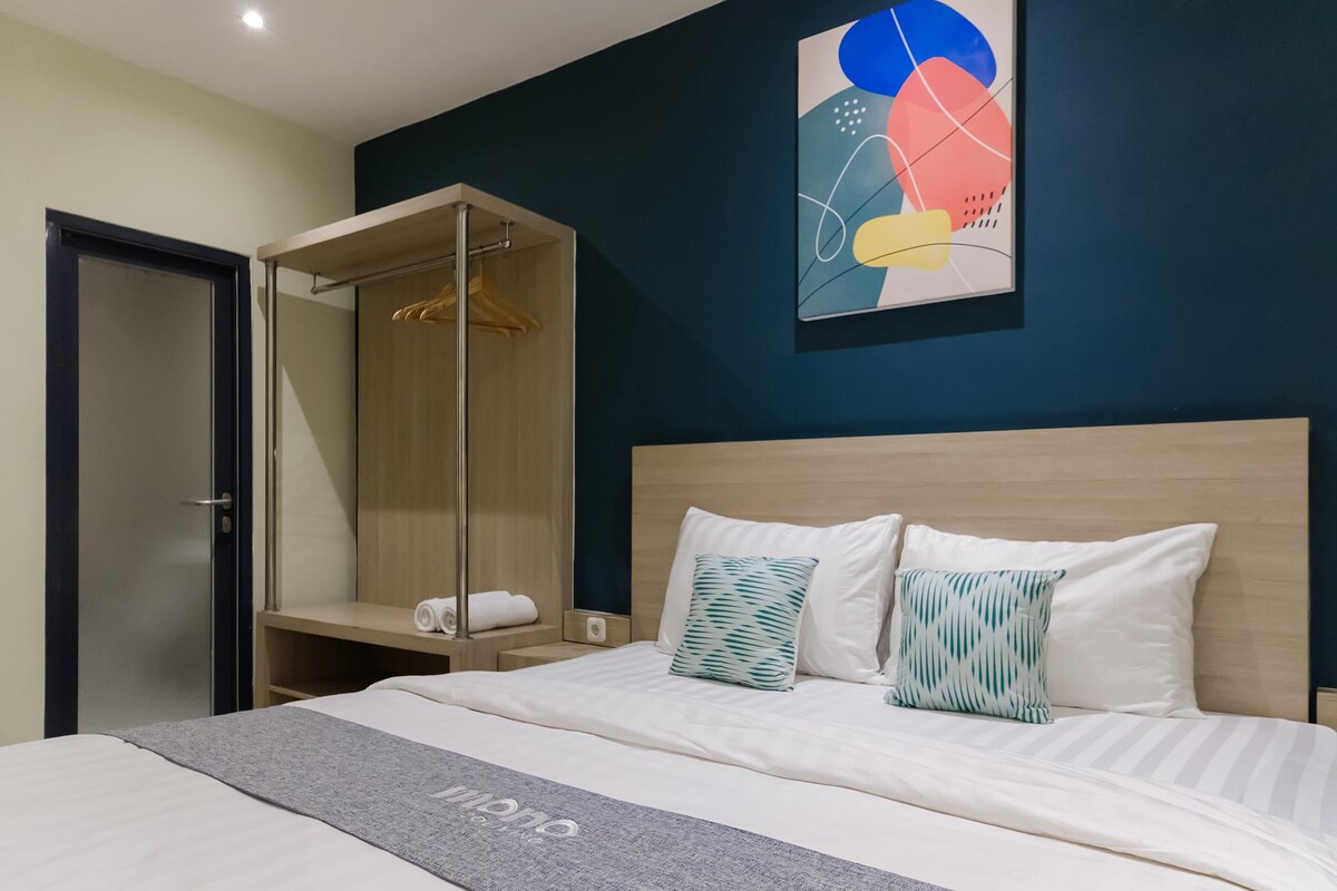 A cozy sleeping area features a spacious bed with light-colored linens, accented by two decorative pillows in shades of blue. A wooden wardrobe is located nearby, displaying hangers and fresh towels. The wall behind the bed is painted dark blue, complemented by colorful abstract art.