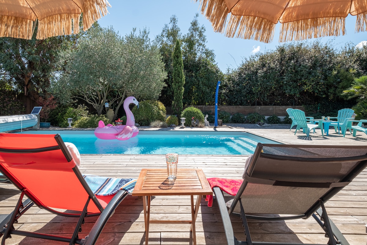 Two lounge chairs are positioned on a wooden deck beside a swimming pool. A small table holds glasses, while a pink inflatable flamingo floats in the pool. Sunny skies and lush greenery create a relaxed outdoor space.