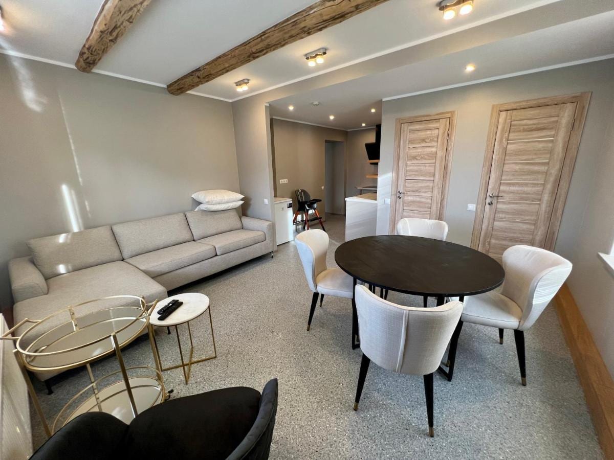 A comfortable living area is shown, featuring a large gray sofa and a round dining table surrounded by four upholstered chairs. Natural light fills the space, highlighting exposed wooden beams above. A small side table and remote are positioned nearby.