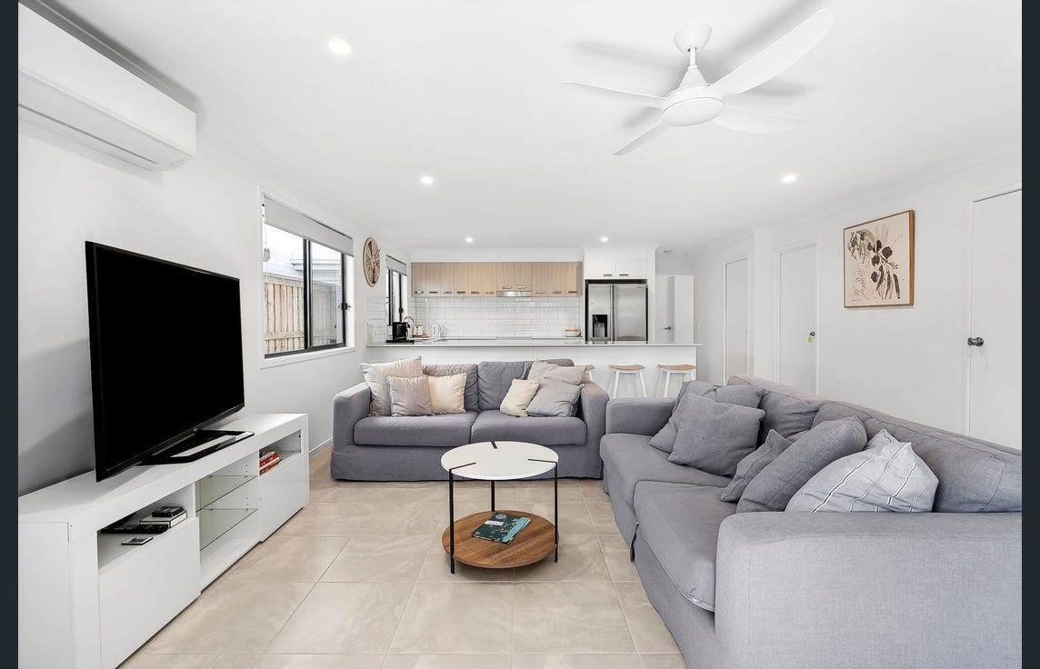 An expansive living area features two large, comfortable sofas arranged around a circular coffee table. A flat-screen TV is positioned opposite the seating. Natural light fills the space through large windows, enhancing the modern, open-concept design.