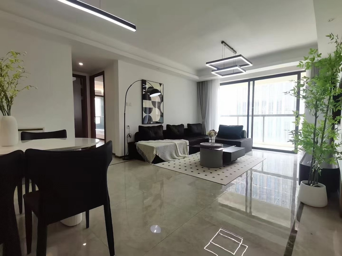 A modern living space features a sleek black sofa complemented by a light-colored throw. A round coffee table sits on a patterned rug, while a dining area is visible in the background. Large windows fill the room with natural light, showcasing the minimalistic decor.