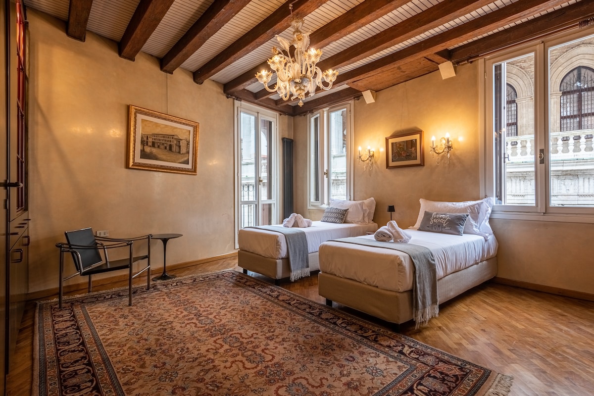 The spacious sleeping area features two beds with light bedding and a rustic wooden ceiling. Elegant chandeliers hang from the ceiling, and large windows provide natural light and views. A classic rug lies on the wooden floor, and a small desk is positioned in the corner.