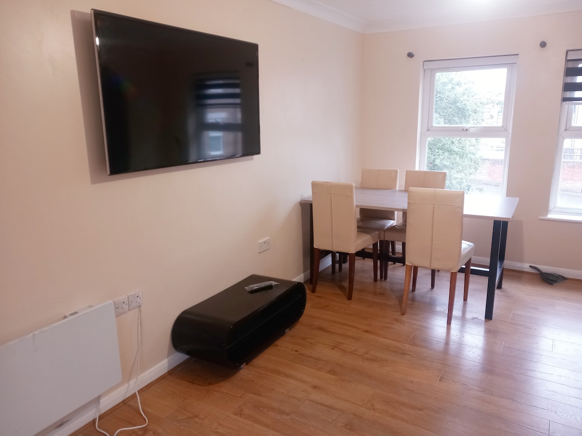 A living area is presented with a wall-mounted flat-screen television above a sleek black entertainment unit. A rectangular dining table with four light-colored chairs is positioned nearby, and two windows allow natural light to enter the space.