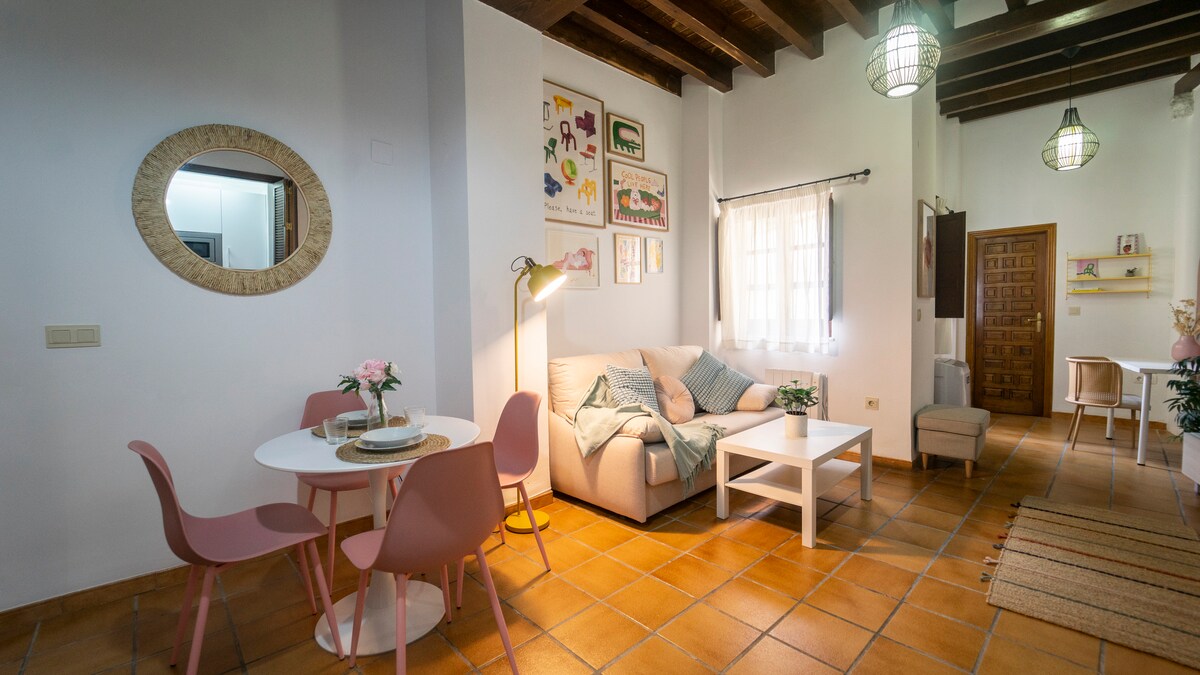 A welcoming living area features a light-colored sofa draped with a soft blanket, complemented by a small round dining table surrounded by pink chairs. Artwork adorns the walls, and natural light filters through a window with sheer curtains, enhancing the cozy ambiance.