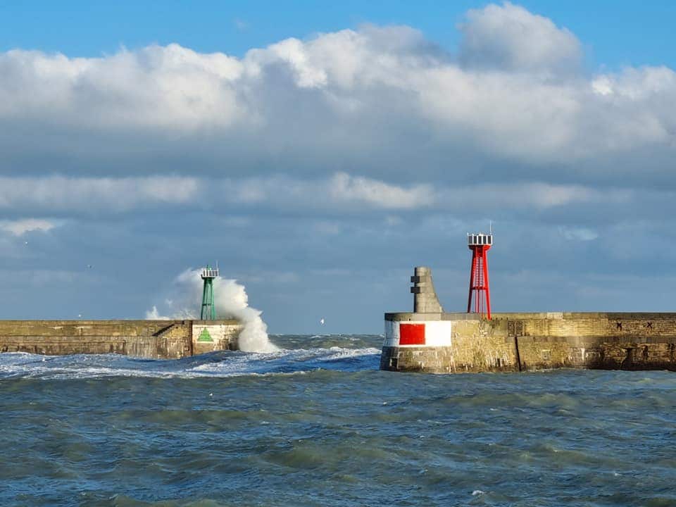 A coastal scene features two contrasting lighthouses positioned at the harbor entrance. One lighthouse is painted red, while the other is green. Waves crash against the stone pier, reflecting dynamic movement under a partly cloudy sky.
