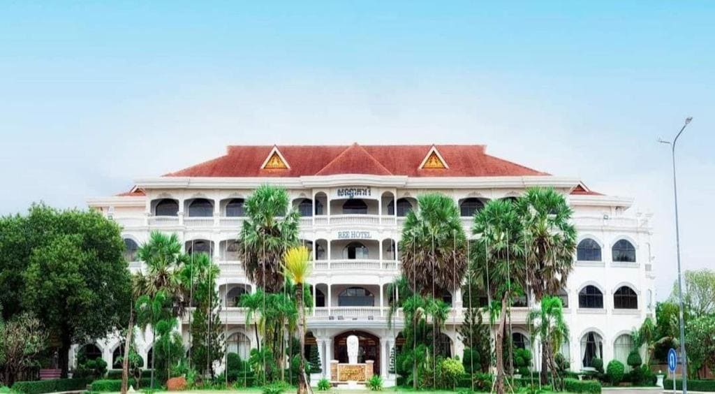A traditional hotel façade is showcased, featuring a three-story structure with a terracotta roof and large windows. Lush green palm trees and well-maintained gardens surround the entrance, creating a welcoming atmosphere. The clear blue sky serves as a backdrop, enhancing the setting.