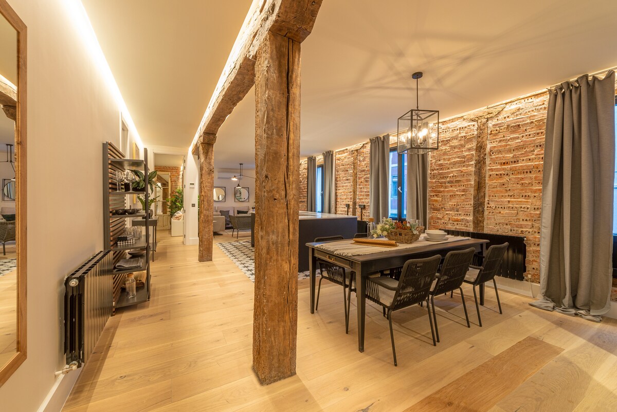 An open layout is showcased, featuring exposed brick walls and wooden beams. A dining area with a long table and chairs is present, complemented by stylish lighting. The flooring is a mix of wood and tiles, while plants add a touch of greenery to the space.