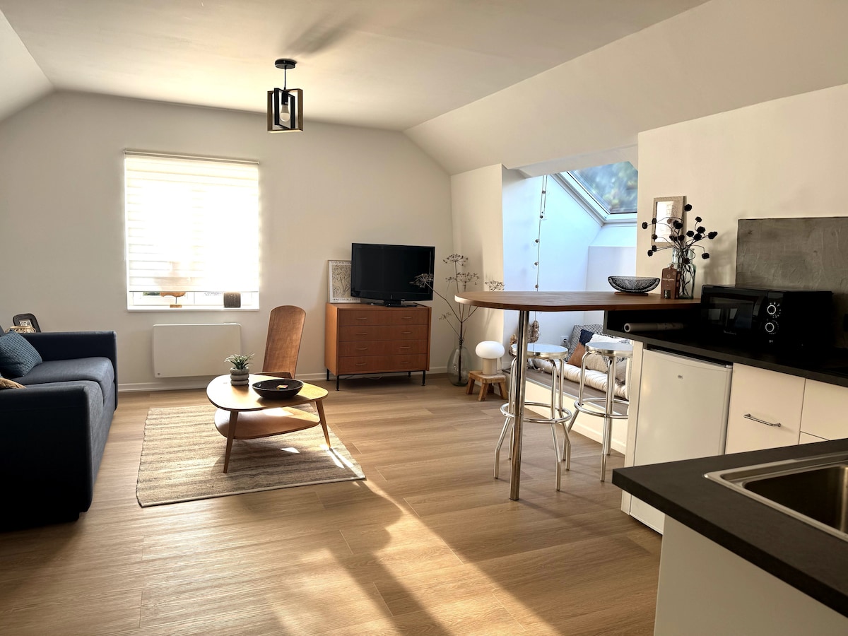 An open living space features a modern sofa and a wooden coffee table. A television is positioned on a low cabinet, with a dining area visible in the background. Natural light floods through a skylight, illuminating the warm-toned flooring.