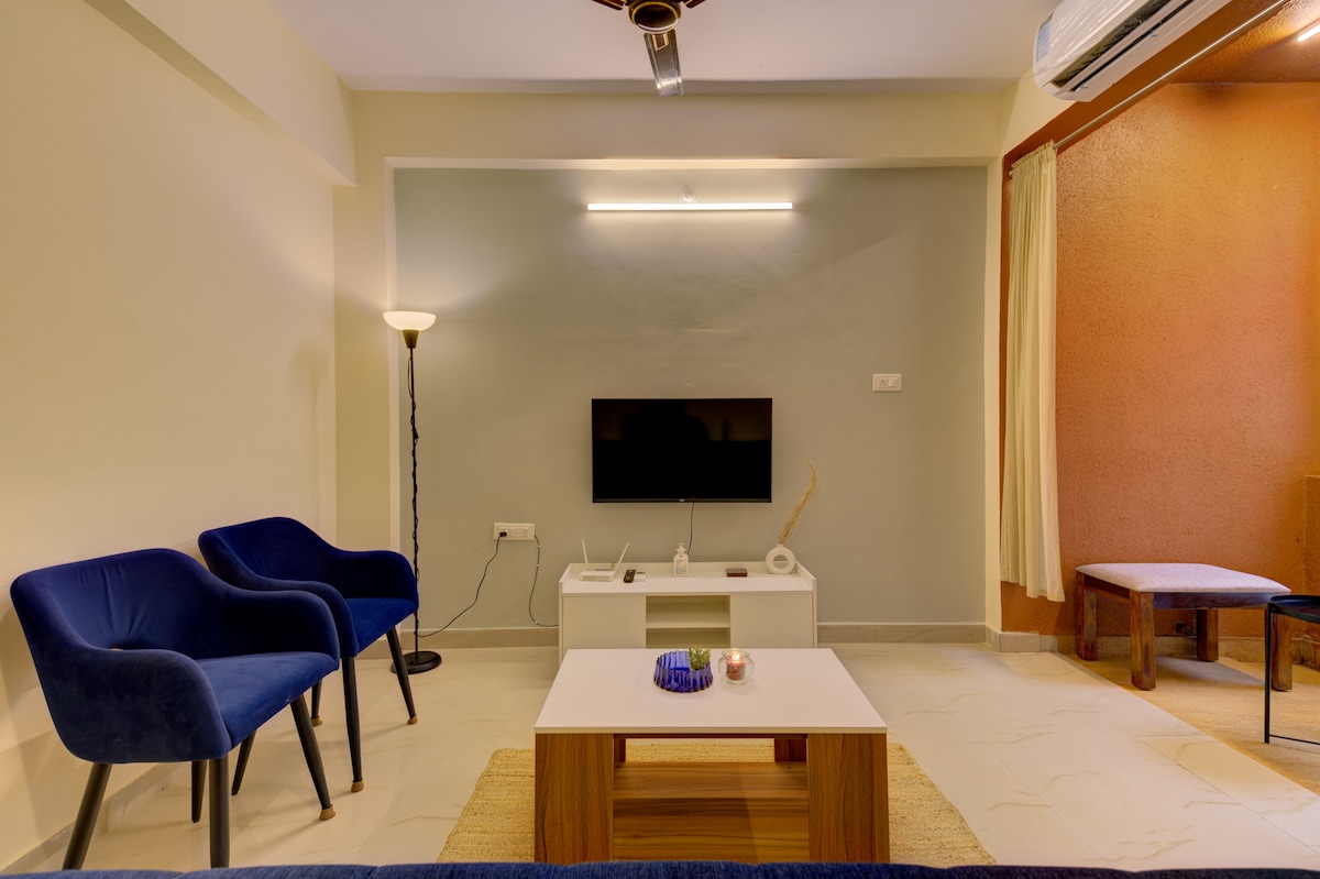 A compact living area features a mounted television against a softly lit wall. Two blue chairs are positioned beside a light wooden coffee table, complemented by a decorative item. A floor lamp stands in one corner, and a small seating area is visible against the opposite wall.