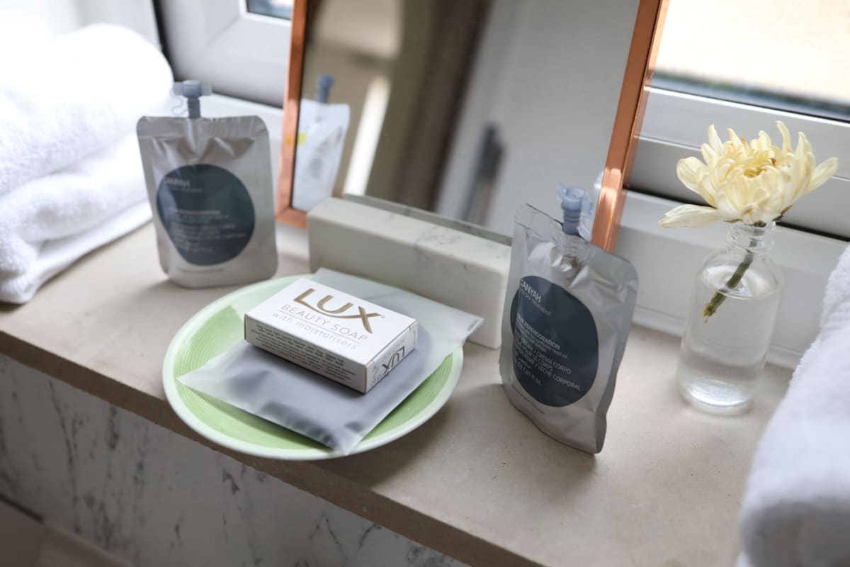 A bathroom sink area is presented, featuring a mirror and a small dish. Luxurious bathing products are displayed, including a bar of soap and two pouches. A vase with a delicate flower adds a touch of elegance, alongside neatly arranged fluffy towels.