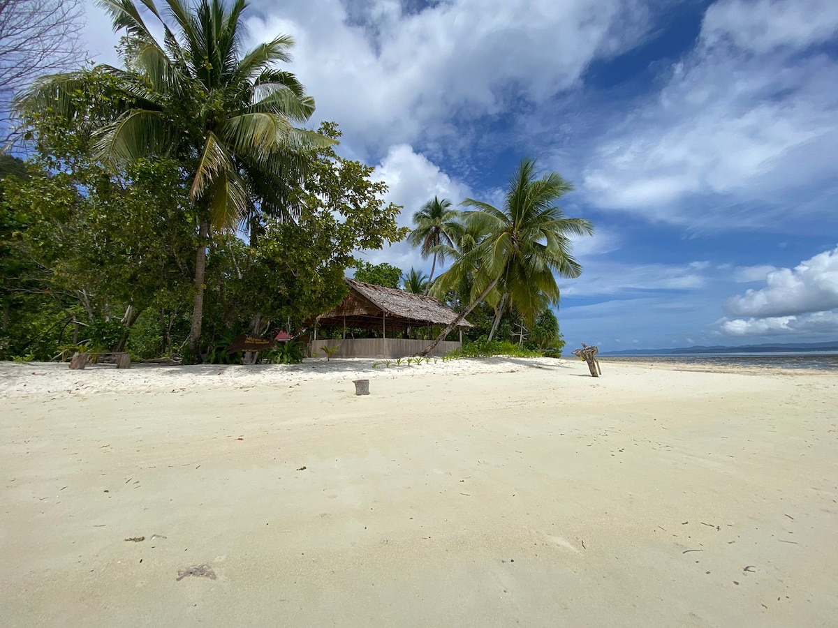 A sandy beach stretches out under a bright sky, with lush green palm trees in the foreground. A traditional wooden structure is partially visible, nestled amid the foliage. The tranquil coastline features clear waters and is bordered by soft, light-colored sand.