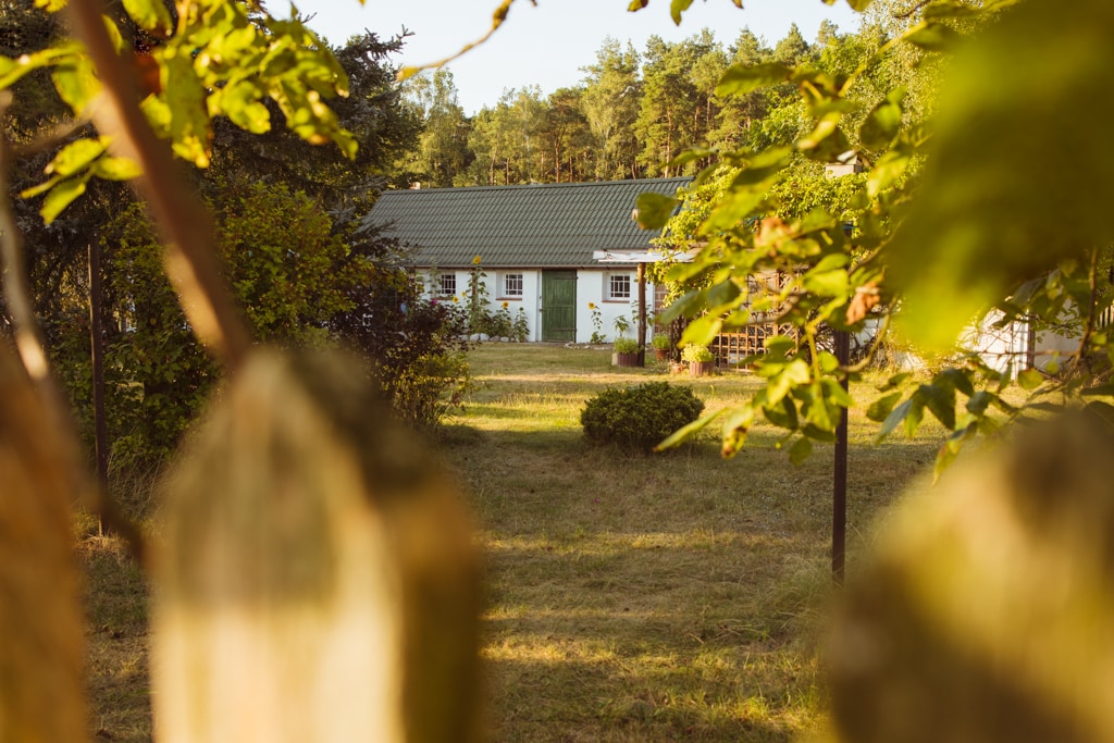 A serene view of a light-colored house is framed by greenery, showing a well-maintained garden. The house features large windows and a welcoming entrance, surrounded by tall trees and open space, contributing to a peaceful outdoor setting.