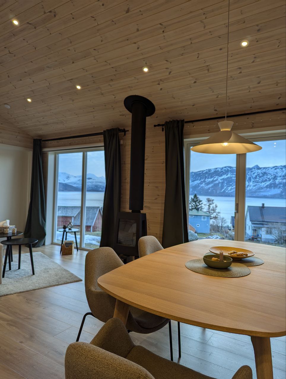 A dining area with a light wooden table is surrounded by gray upholstered chairs. A round pendant light hangs above, illuminating the space. Large glass doors open to a scenic view of mountains and water, while a small seating area with a rug and floor lamp is visible.