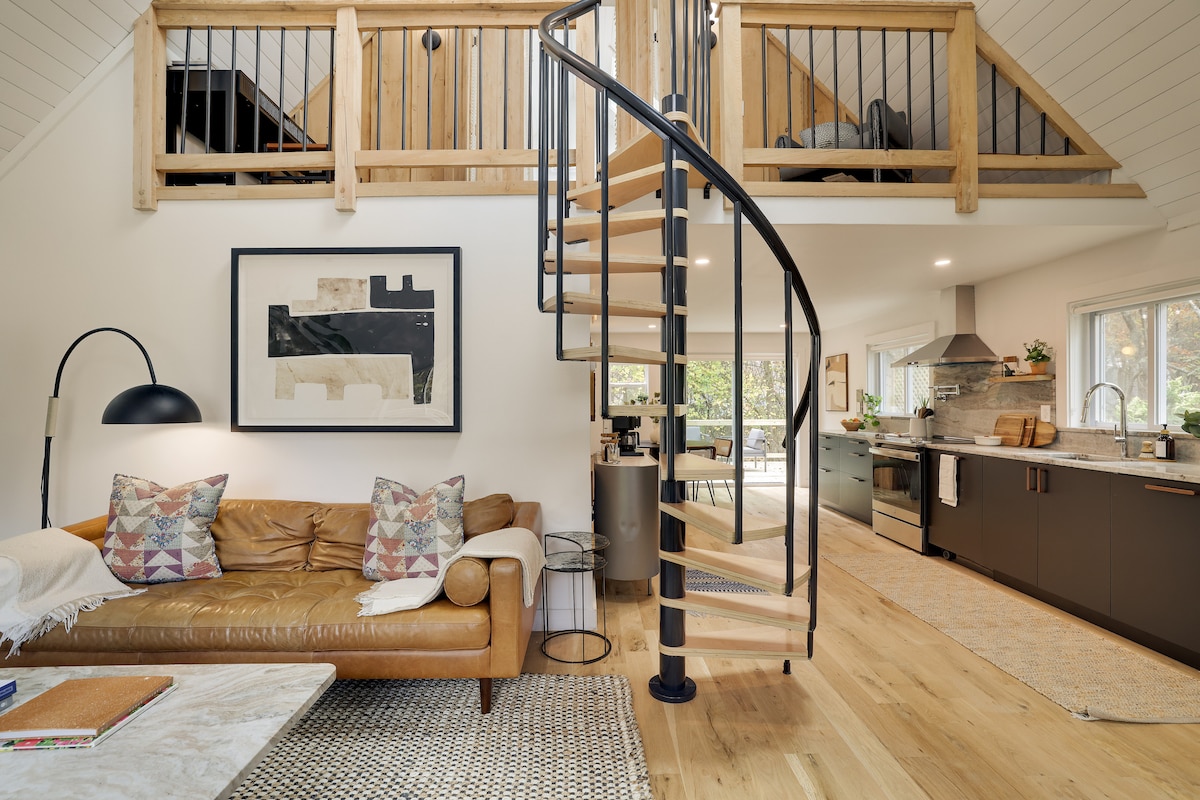 A cozy living area features a brown leather sofa adorned with patterned cushions, positioned next to a round side table. A spiral staircase leads to an upper level, with wooden railings complimenting the open design. Natural light illuminates the space, highlighting the wooden flooring.