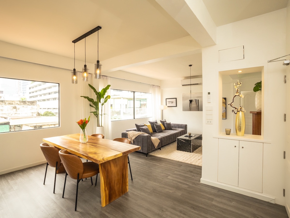 An open living and dining area showcases a large wooden dining table surrounded by four chairs. The space features a cozy seating arrangement with a sofa and decorative cushions. Large windows allow natural light to fill the room, enhancing the modern and airy ambiance.