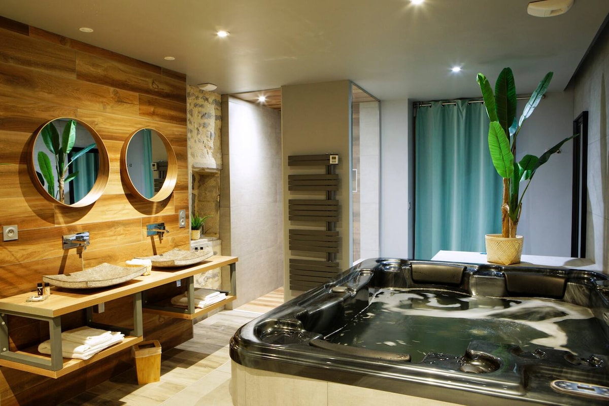 A serene bathroom features a large jacuzzi surrounded by wooden accents, enhancing a relaxing ambiance. Two circular mirrors are mounted above stylish sinks with stone washbasins. A potted plant adds a touch of greenery, while soft lighting creates a calming environment.
