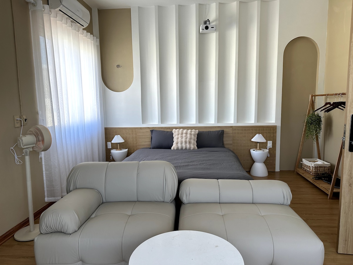 A spacious room features a comfortable bed against a textured wall, complemented by modern bedside tables. A cozy seating area with a light-colored couch invites relaxation. Natural light filters through sheer curtains, enhancing the airy ambiance of the space.