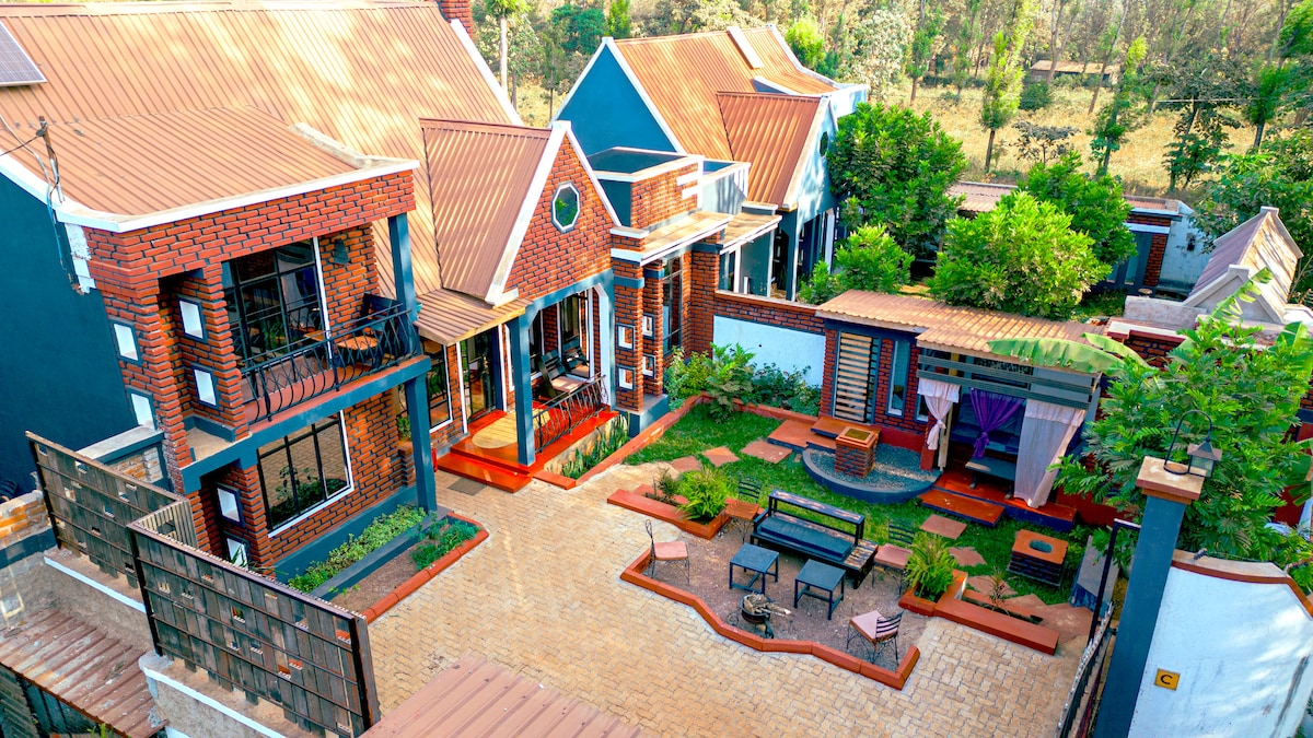 A spacious outdoor area features a mix of brick and blue houses set among greenery. The courtyard has various seating arrangements and a shaded area, providing an inviting space for gathering. Lush trees and plants enhance the natural surroundings, creating a serene environment.