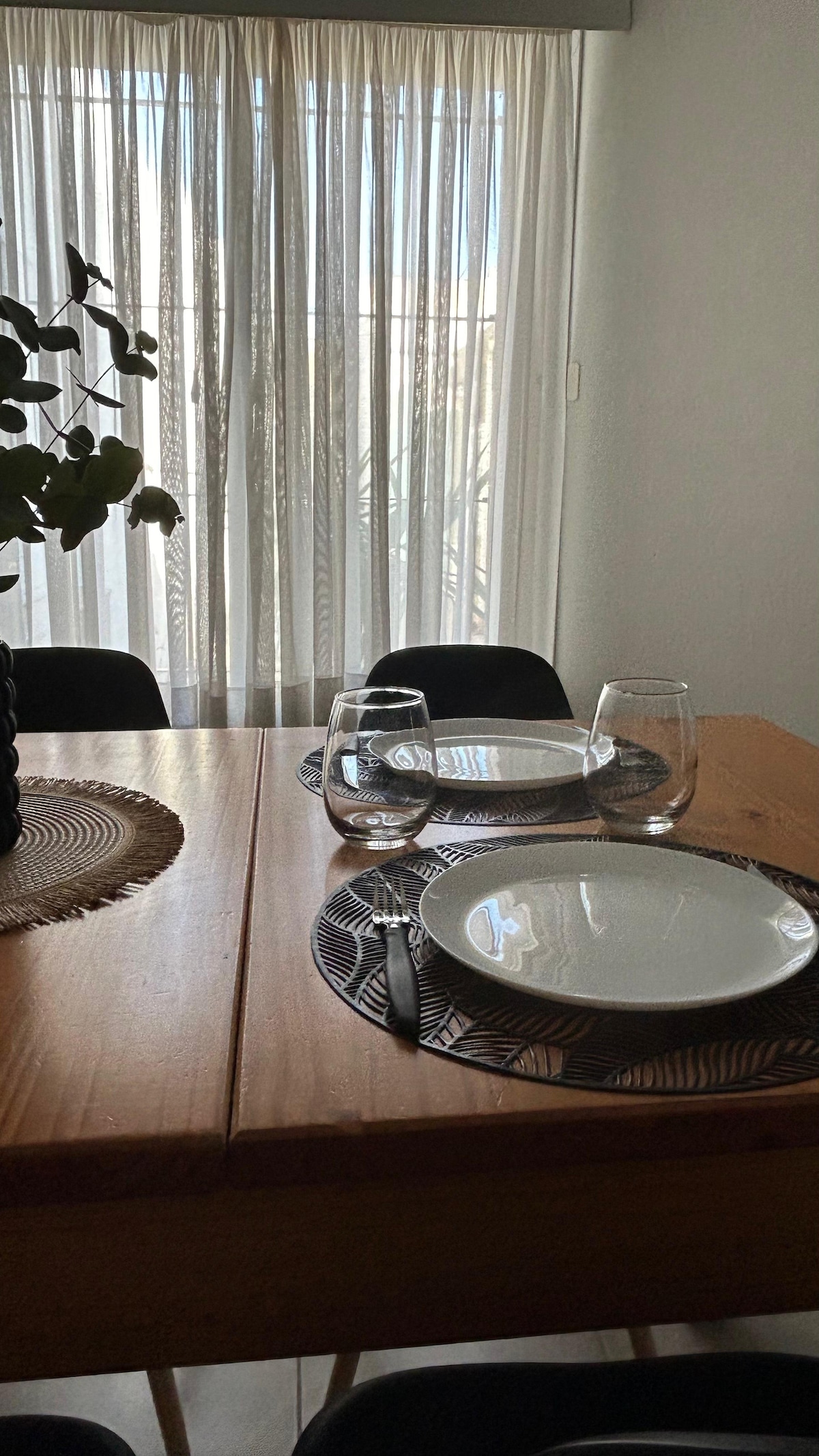 A wooden dining table is set with two plates and glasses, complemented by a woven placemat. A large window covered with sheer curtains allows natural light to fill the space, creating a cozy ambiance. A plant is visible in the background.