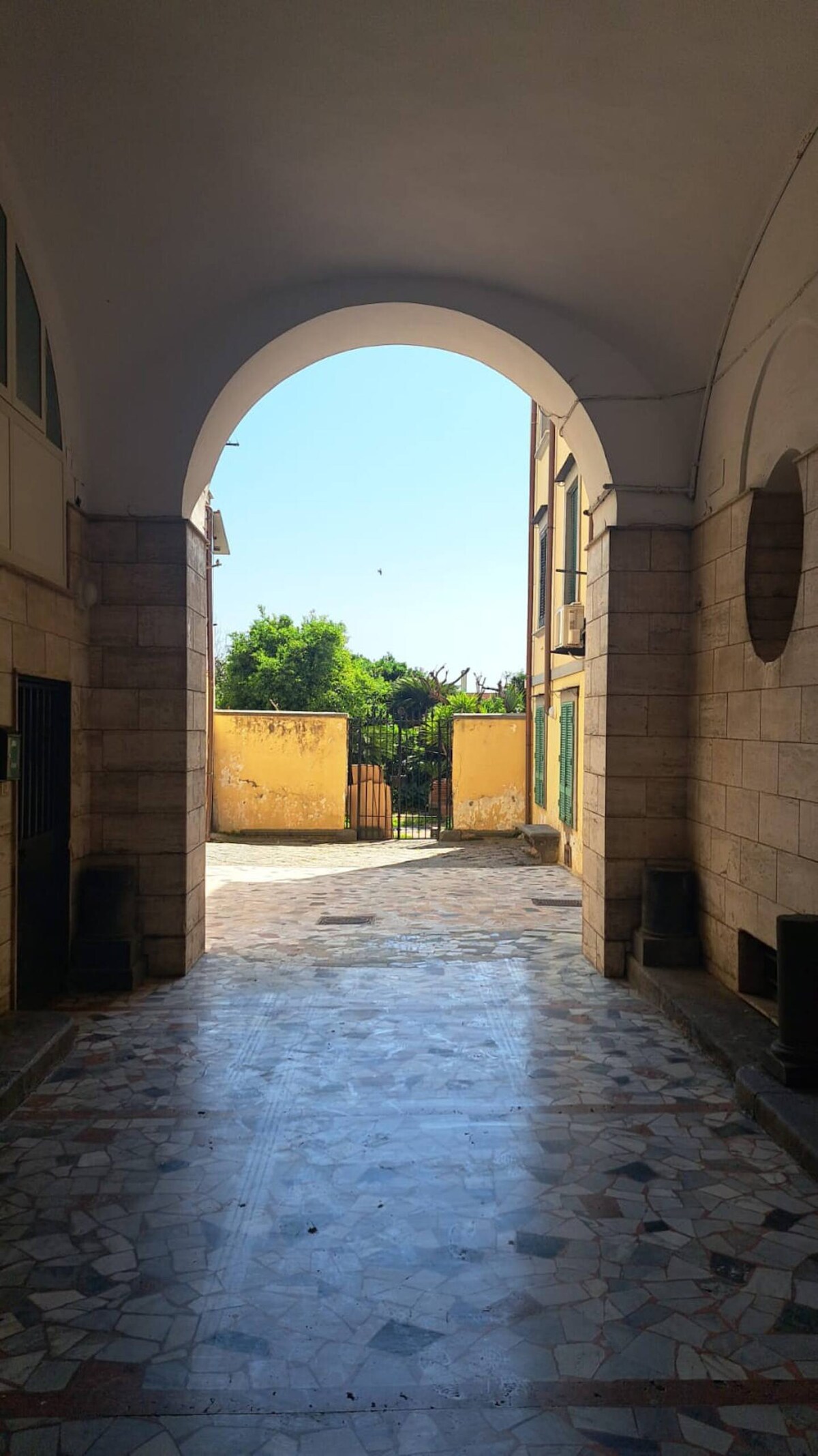 An arched entrance is shown, leading to an outdoor space with lush greenery visible in the background. The smooth, patterned floor extends towards a gate framed by yellow walls, creating an inviting transition from the interior passage to the exterior area.