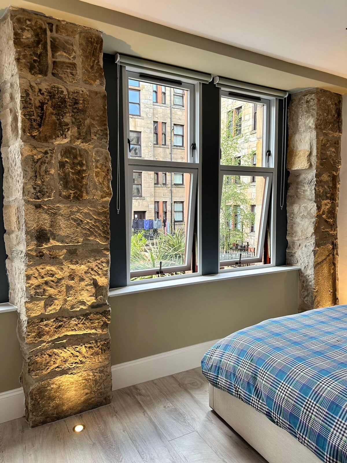 Natural stone walls frame the large windows, which offer views of the outdoor greenery and neighborhood. A checkered blanket covers the spacious bed beneath the windows, with soft lighting accentuating the textures of the room.