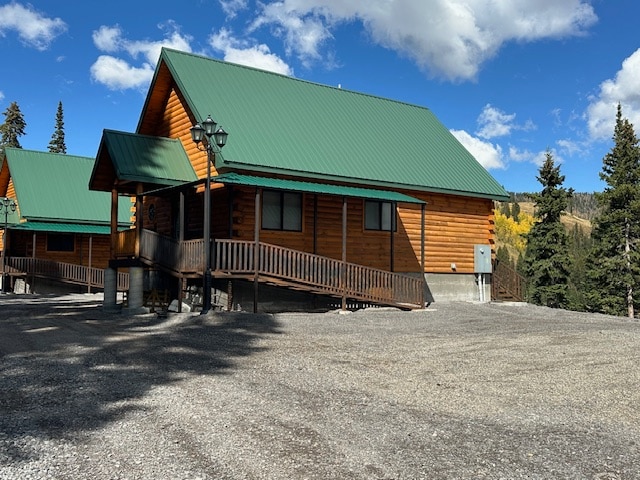 A charming cabin features a sloped green metal roof and natural wood siding. A wooden ramp leads to the entrance, surrounded by gravel and tall trees. Soft sunlight highlights the structure against a backdrop of blue skies and scattered clouds.