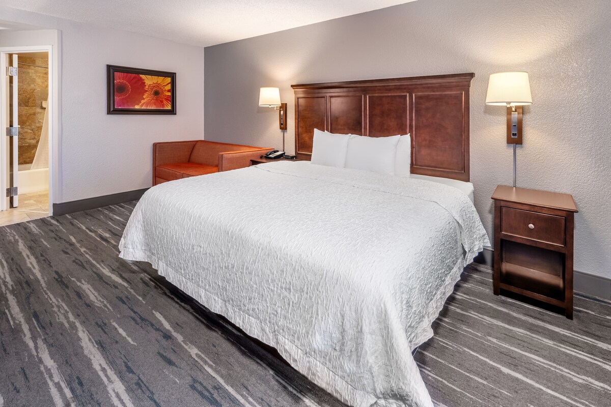 A spacious hotel room features a large bed with crisp white linens, complemented by a wooden headboard. A wooden nightstand, containing a lamp, is positioned beside the bed. An orange sofa is situated against the wall, and a colorful artwork adorns the wall.