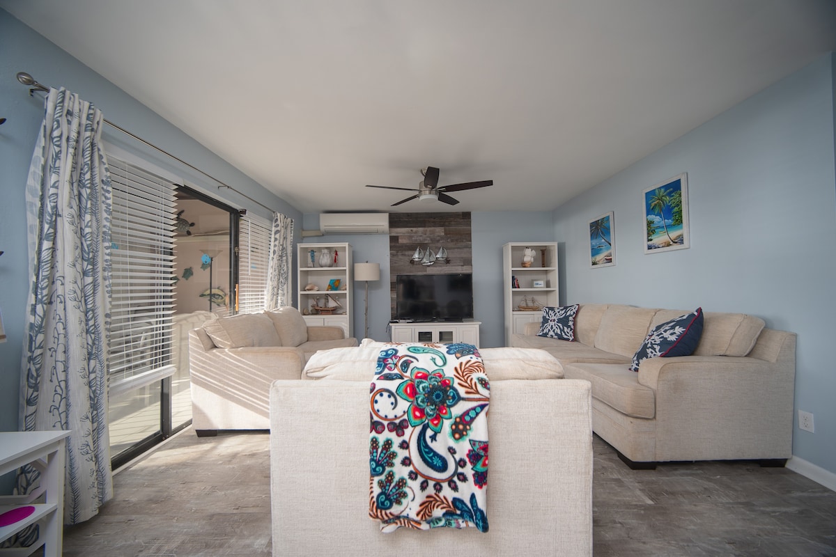 A bright living area is presented with soft blue walls and natural light filtering through large sliding doors. Comfortable seating options include two light-colored sofas and decorative cushions. Shelves are arranged with decorative items, while framed beach art adorns the walls.