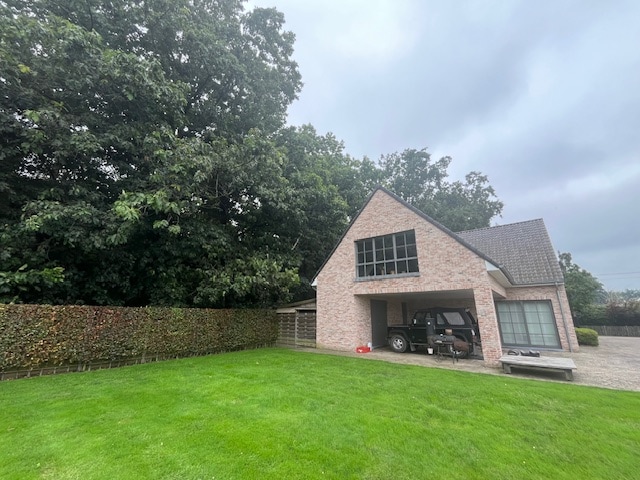 The exterior of the house is showcased, featuring a brick facade and large windows. A well-maintained lawn extends in front, bordered by lush greenery and hedges. A vehicle is parked in the driveway, highlighting the convenient access to the property.