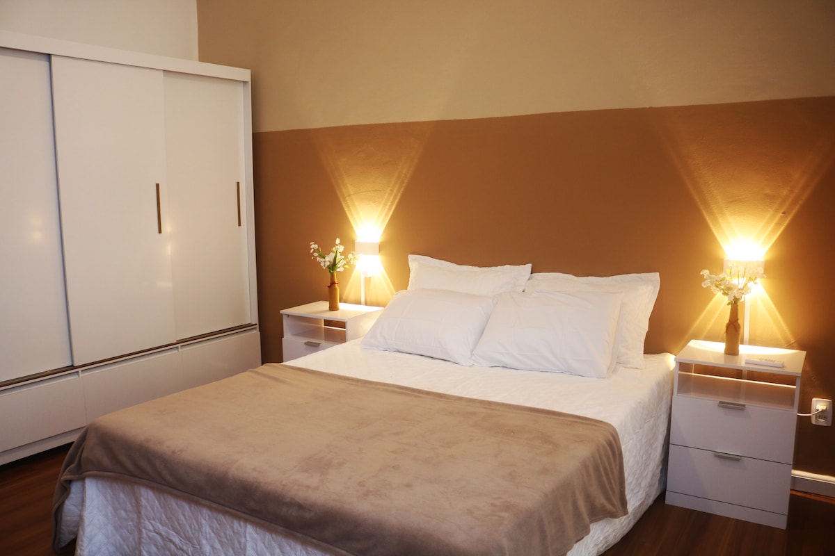 A neutral-toned bedroom features a spacious bed dressed in white linens and a light beige throw. Flanking the bed are two bedside tables with soft glowing lamps. A large wardrobe is positioned against the wall, contributing to the room's organized appearance.
