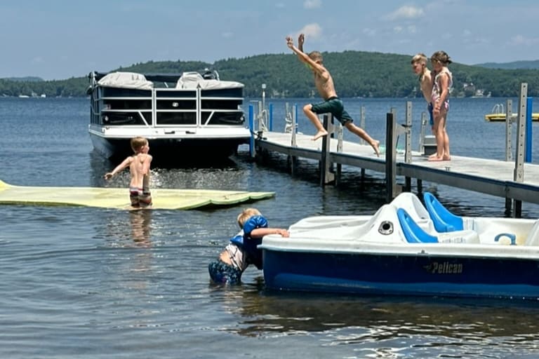 A vibrant scene captures children playing in the water near a dock. One child is diving off the dock, while another leans over a small paddle boat. Inflatable float mats rest on the surface, alongside surrounded by a serene lake and lush green hills in the background.